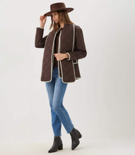 Load image into Gallery viewer, A woman modeling a dark brown quilted jacket with white trim, blue jeans, black ankle boots, and a brown wide-brim hat against a plain white studio background.
