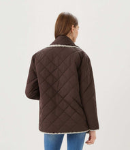 Load image into Gallery viewer, A woman viewed from the back wearing a dark brown diamond-quilted jacket with cream-colored trim along the collar and hem, paired with blue jeans, against a plain white studio background.
