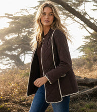 Load image into Gallery viewer, A woman wearing a dark brown quilted jacket with cream-colored scalloped trim, standing outdoors in a natural setting with trees and dry grass in the background.
