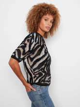 Load image into Gallery viewer, A woman with curly brown hair wearing a short-sleeve black and beige zebra-pattern knit top and blue jeans, posing against a light gray studio background.
