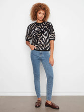 Load image into Gallery viewer, A woman modeling a short-sleeve knit top with a black and beige zebra-like pattern, paired with blue skinny jeans and brown loafers, standing against a plain white wall.

