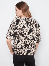 Load image into Gallery viewer, A woman shown from the back wearing a short-sleeve top with a black, brown and off-white cow print pattern, paired with black jeans, against a plain light gray background.
