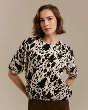 Load image into Gallery viewer, A woman wearing a short-sleeve knit top with a black, brown and off-white cow-print pattern, paired with dark brown pants, standing against a solid beige studio background.
