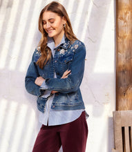Load image into Gallery viewer, A woman wearing a blue denim jacket with floral embroidery and rhinestones on the shoulders, layered over a blue and white striped button-down shirt and paired with burgundy pants, posing against a textured white wall with natural shadows.
