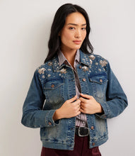 Load image into Gallery viewer, A woman wearing a blue denim jacket with floral embroidery and sequins on the chest, layered over a striped button-down shirt and burgundy pants, photographed against a plain light gray background.
