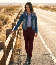 Load image into Gallery viewer, A woman walks down a desert road wearing a blue denim jacket with floral embroidery and rhinestones on the shoulders, a light blue button-up shirt, burgundy pants, and black boots.
