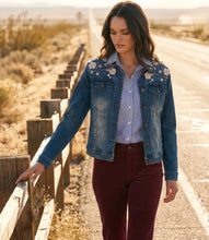 Load image into Gallery viewer, A woman stands on a paved road beside a wooden fence in a desert landscape, wearing a blue denim jacket with white floral embroidery and rhinestones on the shoulders over a blue and white striped button-down shirt and burgundy pants.
