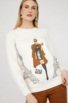 Woman wearing a sweater with a graphic design of a woman in a coat and Eiffel Tower and brown pants against a white background.