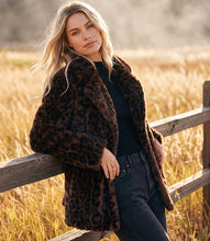 Load image into Gallery viewer, A woman leans against a wooden fence in a sunlit field, wearing a dark brown leopard-print faux fur coat over a black top and dark jeans.
