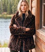 Load image into Gallery viewer, A woman wearing a brown and black leopard-print faux fur coat stands with arms crossed next to a rustic wooden cabin, with a lake and pine forest in the background.
