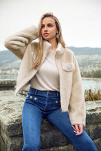 Load image into Gallery viewer, Woman wearing a champagne faux fur jacket and blue jeans sitting outdoors with mountains in the background.
