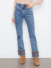 Load image into Gallery viewer, A person wearing medium-wash blue jeans with decorative embroidered trim at the hem, paired with a beige ribbed top and tan ankle boots, standing against a white wall.
