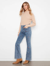 Load image into Gallery viewer, A woman modeling a beige ribbed mock-neck long-sleeve top and high-waisted blue flare jeans with embroidered hearts, paired with tan ankle boots, against a white wall.
