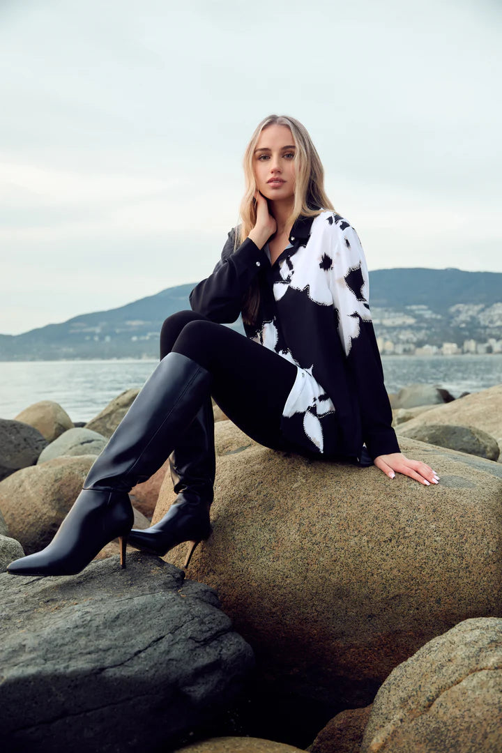 Woman sitting on rocks by a body of water wearing a black and white floral tunic blouse, black leggings and black tall boots.
