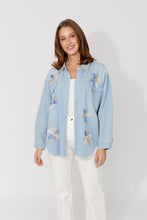 Load image into Gallery viewer, Woman wearing a light blue denim shirt with floral embroidery over a white top and white pants on a white background.
