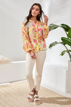 Load image into Gallery viewer, Woman wearing a colorful blouse, white pants and white sandals standing in a bright room with a plant.
