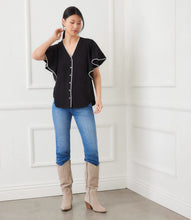 Load image into Gallery viewer, Woman wearing a black short sleeve blouse with cream trim, blue jeans, and beige boots against a white wall.
