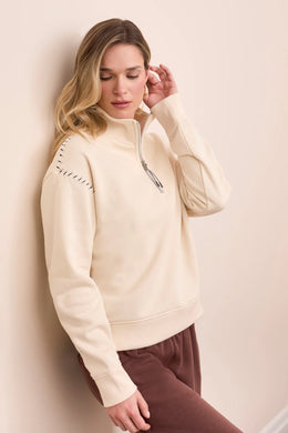 Woman wearing a moonstone zip-up sweater and brown pants standing against a beige wall.