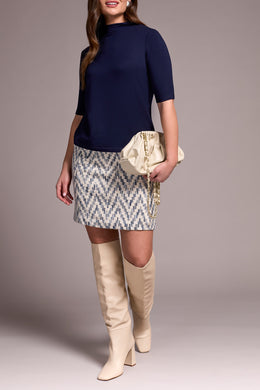 Woman wearing a navy top, patterned skirt, and beige knee-high boots with a beige clutch against a neutral background.