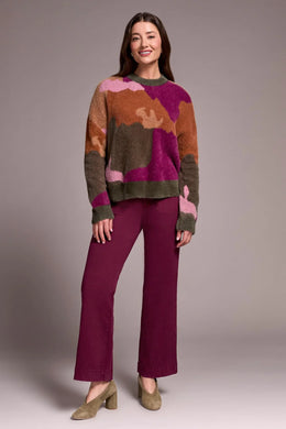 Woman wearing a colorful abstract print sweater, purple pants and olive suede heel shoes against a plain background.