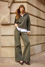 Load image into Gallery viewer, Woman wearing a white blouse, avocado green sweater, avocado green wide leg denim and heels standing against a stone wall.
