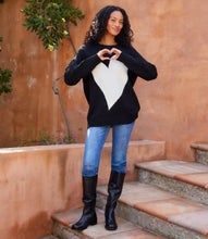 Load image into Gallery viewer, Woman wearing a black and white heart-patterned sweater and blue jeans, standing on steps with a warm-toned wall and plant in the background.
