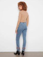 Load image into Gallery viewer, A back view of a woman with curly hair stands against a white wall wearing a brown long-sleeve top, blue jeans with floral embroidery at the hem and black boots.

