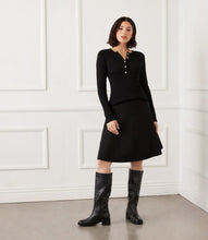 Load image into Gallery viewer, Woman wearing a black ribbed sweater with rhinestone buttons, black skirt and tall black boots against a white wall.
