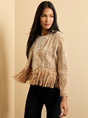 Woman wearing a beige fringed jacket, white top and black pants against a beige background.