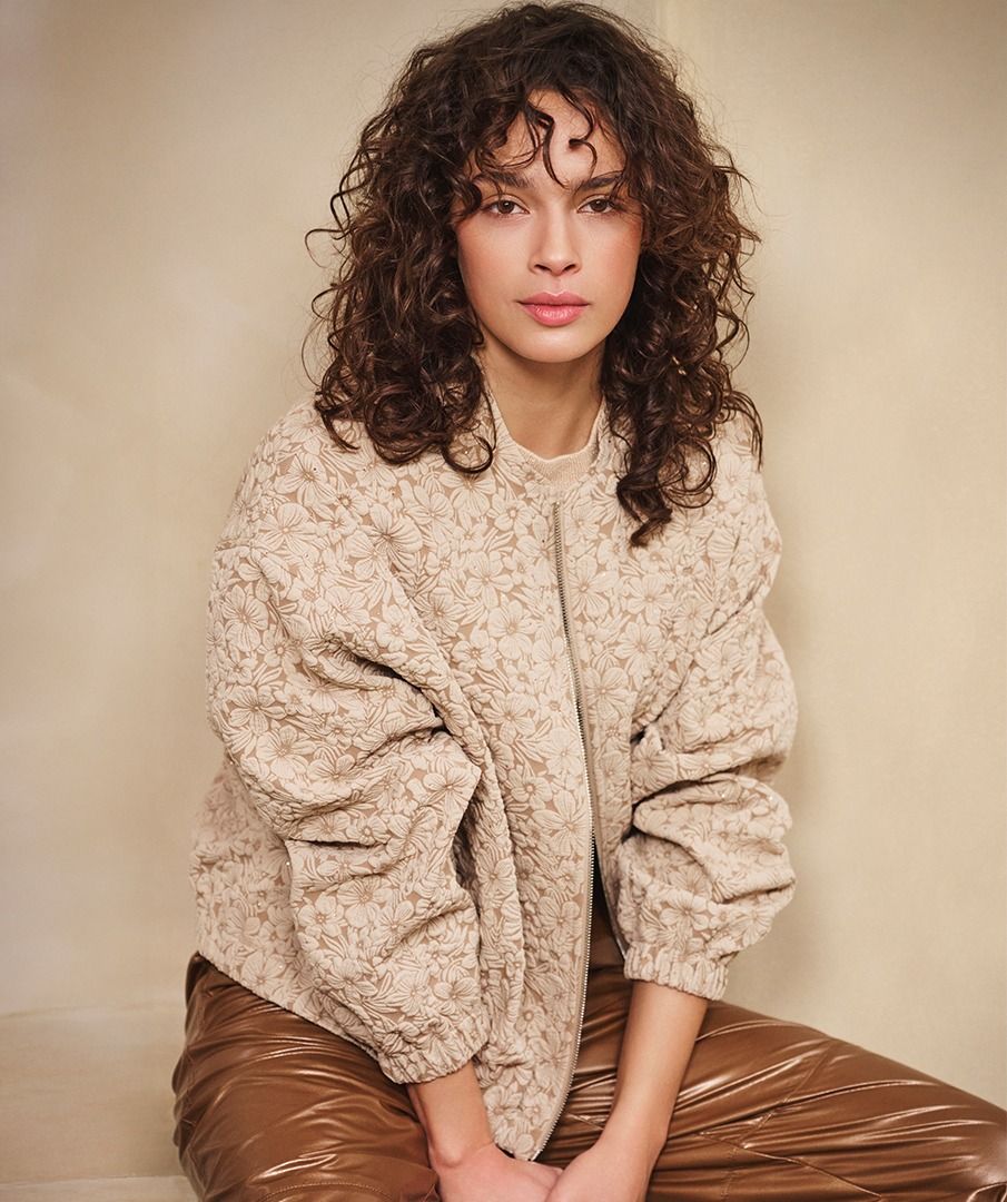 Woman wearing a beige quilted jacket and brown pants against a beige background.