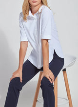 Load image into Gallery viewer, A woman seated on a white and wood stool wearing a white short-sleeve button-up shirt and dark navy pants, photographed against a light neutral studio background.
