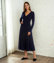 Load image into Gallery viewer, Woman wearing a navy blue dress standing in a room with white brick walls and a door.
