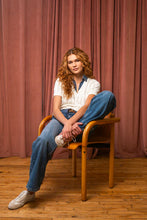 Load image into Gallery viewer, Woman wearing a white knitted and denim collared and hem short sleeve top, blue jeans and sneakers sitting in a chair in front of a brown curtain.
