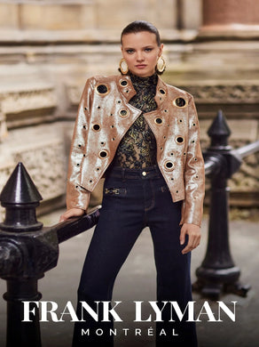 Model wearing a navy and gold floral lace top with a gold jacket and denim pants standing against a black iron barrier in front of a stone building.