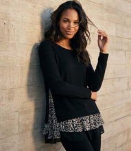 Load image into Gallery viewer, A woman wearing a black long-sleeve top with a black and white leopard print ruffle hem, leaning against a light-colored textured concrete wall.
