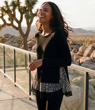 Load image into Gallery viewer, A woman wearing a black long-sleeve knit top with a leopard-print ruffle hem, standing outdoors on a balcony with a glass railing and desert scenery in the background.
