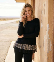 Load image into Gallery viewer, A woman stands outdoors against a weathered tan wall, wearing a black long-sleeve top with a leopard-print ruffle hem and black pants.
