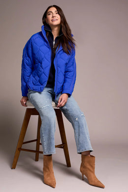 Woman wearing a blue puffer jacket , blue jeans and tan suede boots sitting on a wooden stool against a plain background.
