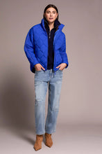 Load image into Gallery viewer, Woman wearing a blue puffer jacket, blue jeans and tan suede boots against a neutral background.

