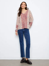 Load image into Gallery viewer, A woman stands against a white wall wearing a loose, long-sleeve knit sweater with a multicolor pattern  paired with dark blue jeans and dark brown loafers.
