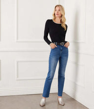Load image into Gallery viewer, Woman wearing a black top with white trim, blue jeans, a black belt and silver shoes against a white wall.
