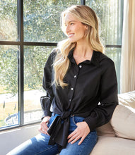 Load image into Gallery viewer, Woman wearing a long sleeve black shirt with a tie waist and blue jeans sitting on a couch in front of large windows with a view of a yard.
