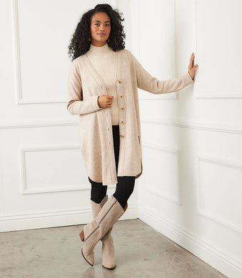 Woman wearing a long beige cardigan over a beige turtleneck, black pants, and tall beige boots against a white wall.