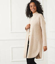 Load image into Gallery viewer, Woman wearing a long beige cardigan over a beige turtleneck and black pants against a white wall.

