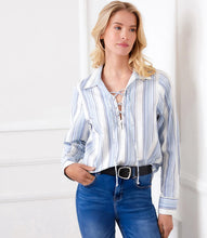 Load image into Gallery viewer, Woman wearing a blue and white striped shirt with a lace-up front, blue jeans and black belt, standing against a white wall.
