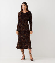 Load image into Gallery viewer, A woman modeling a long-sleeve midi dress in dark brown velvet with an intricate floral pattern, paired with black lace-up strappy sandals against a plain light gray studio background.
