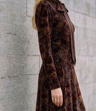 Load image into Gallery viewer, A woman wearing a long-sleeve brown velvet dress with an intricate paisley pattern in dark blue and gold, featuring a matching brown satin bow at the collar, standing against a textured gray concrete wall.
