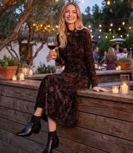 Load image into Gallery viewer, A woman sits on a wooden deck railing at dusk, wearing a dark brown velvet long-sleeve midi dress and black ankle boots, holding a glass of red wine.
