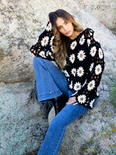 Load image into Gallery viewer, A woman sitting on a large rock outdoors, wearing a black knit sweater with a white daisy floral pattern and blue jeans.
