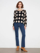 Load image into Gallery viewer, A woman modeling a black long-sleeve crochet top with a white daisy pattern, paired with blue jeans and tan ankle boots, standing against a plain white wall.
